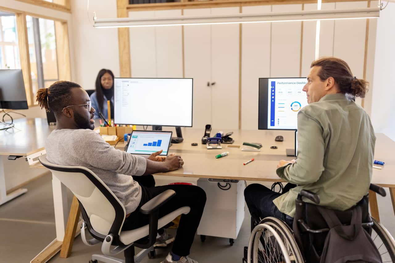 Business colleagues discussing work in office, a man sitting in an office chair next to another in a wheelchair at their desks.