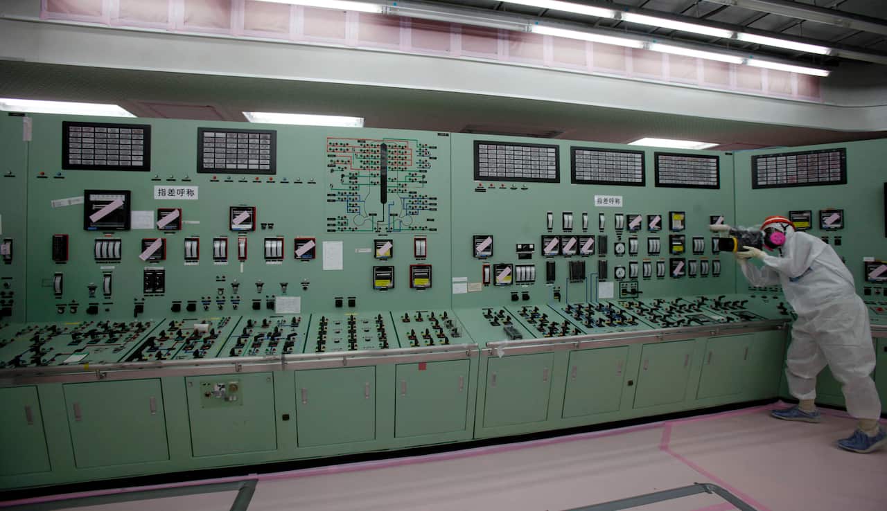 Someone dressed in protective gear takes a photo next to a green panel of switches