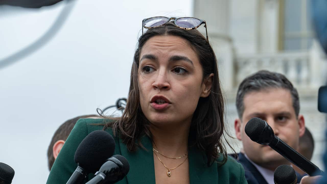 Alexandria Ocasio-Cortez speaking into several black microphones being held up near her.