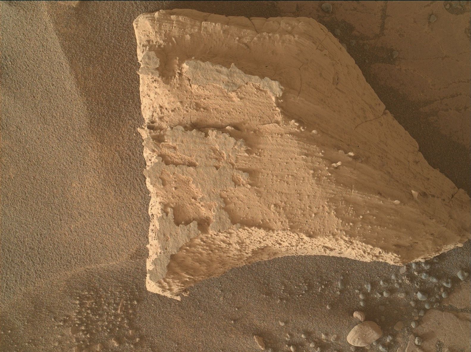 A close-up color photo from the Martian surface shows an area of pale orange, fine-grained soil, resembling a sheet of sandpaper. A large trapezoid-shaped stone sits atop that, filling most of the right two-thirds of the frame. The top surface of the stone facing the viewer is uneven, with numerous lines running left to right, suggesting it’s made of many layers.