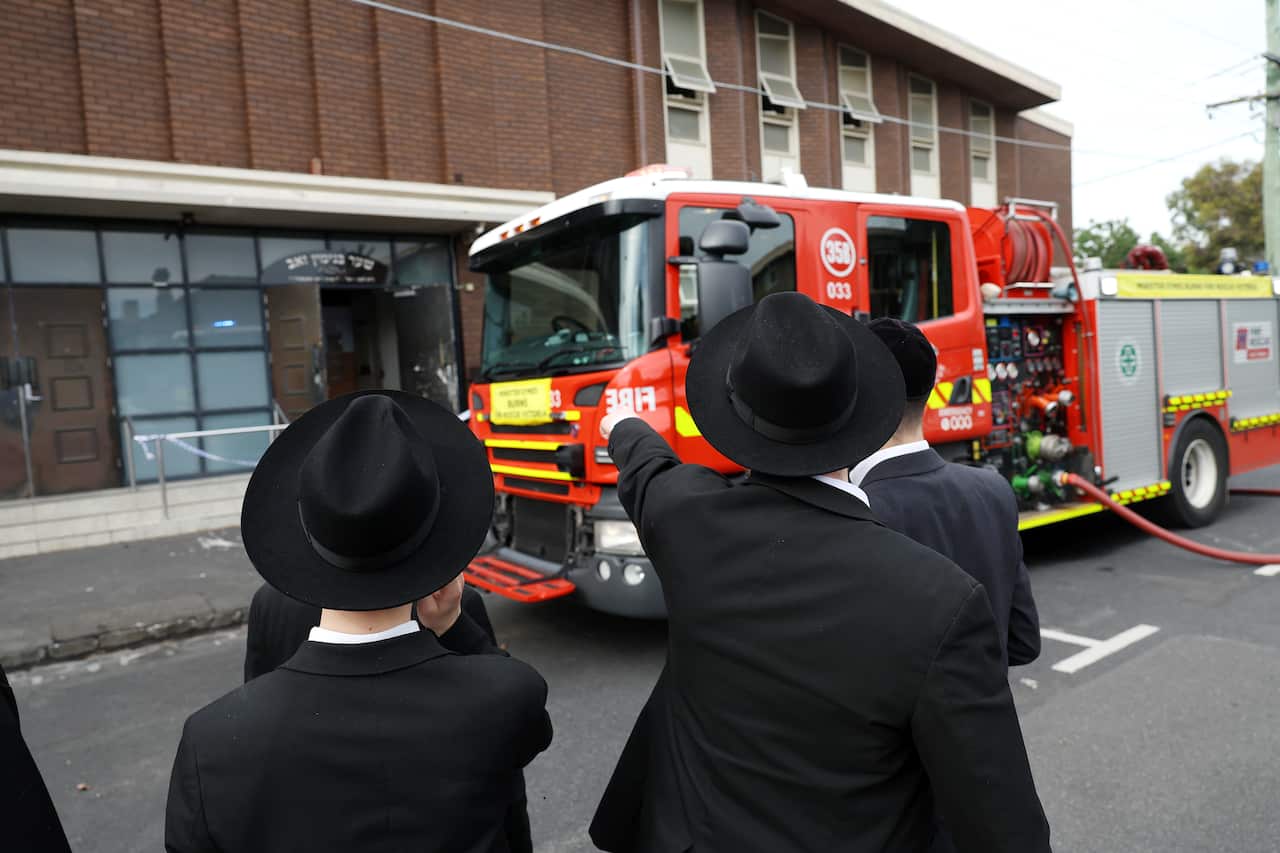 People dressed in black coats and matching hats point at a building in front of which a fire truck is parked.