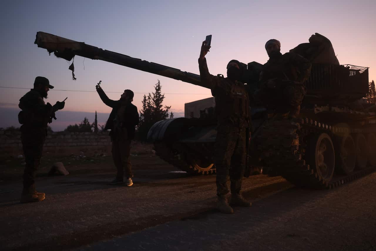 Silhouetted people wearing military equipment stand near a tank on a road.
