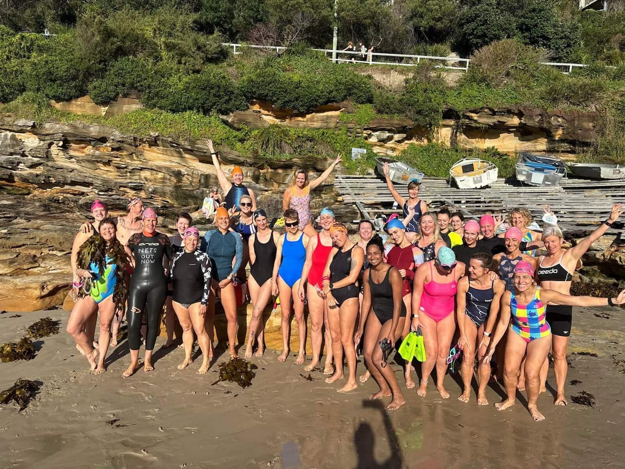 A group in multicoloured bathers and black wetsuits stand before a rocky cliff and some boats. 