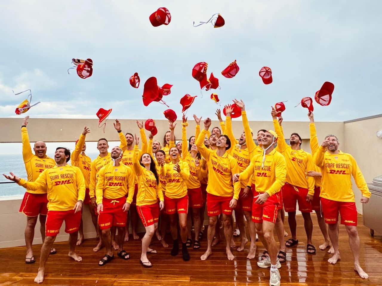 A group of surf life savers in yellow and red patrol uniforms throw their red caps into the air.