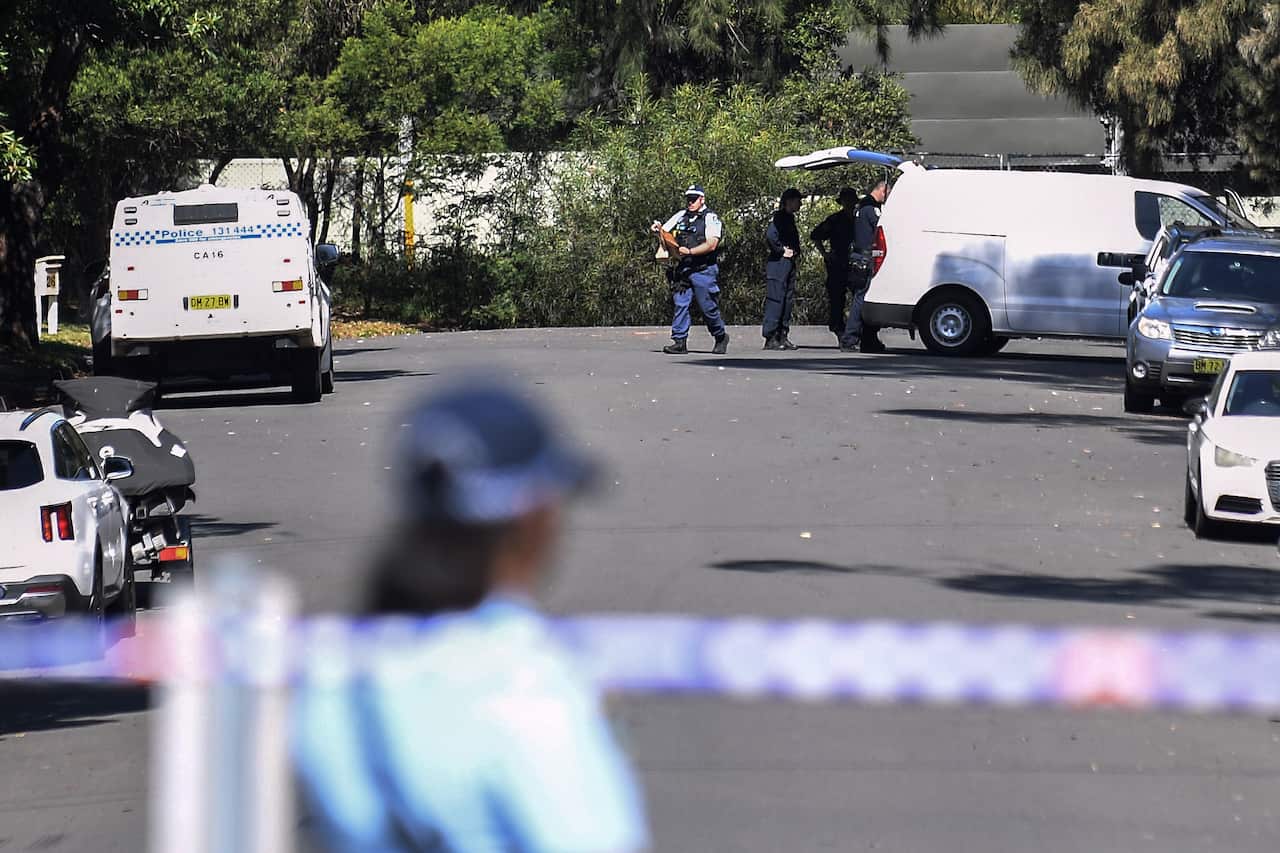 An out-of-focus police officer and police tape in the foreground, with police vans and officers in the background.