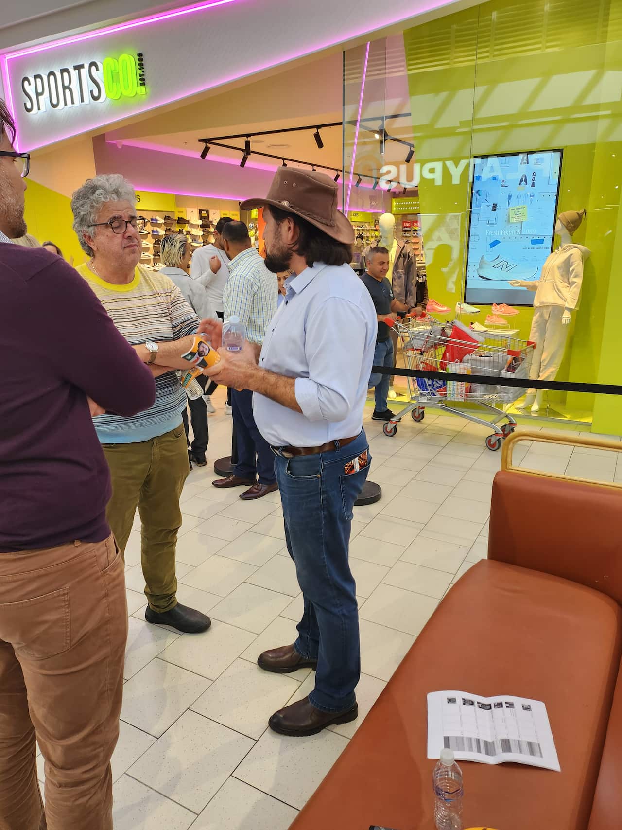 A man in a cowboy hat and jeans is speaking to two men in a shopping centre.