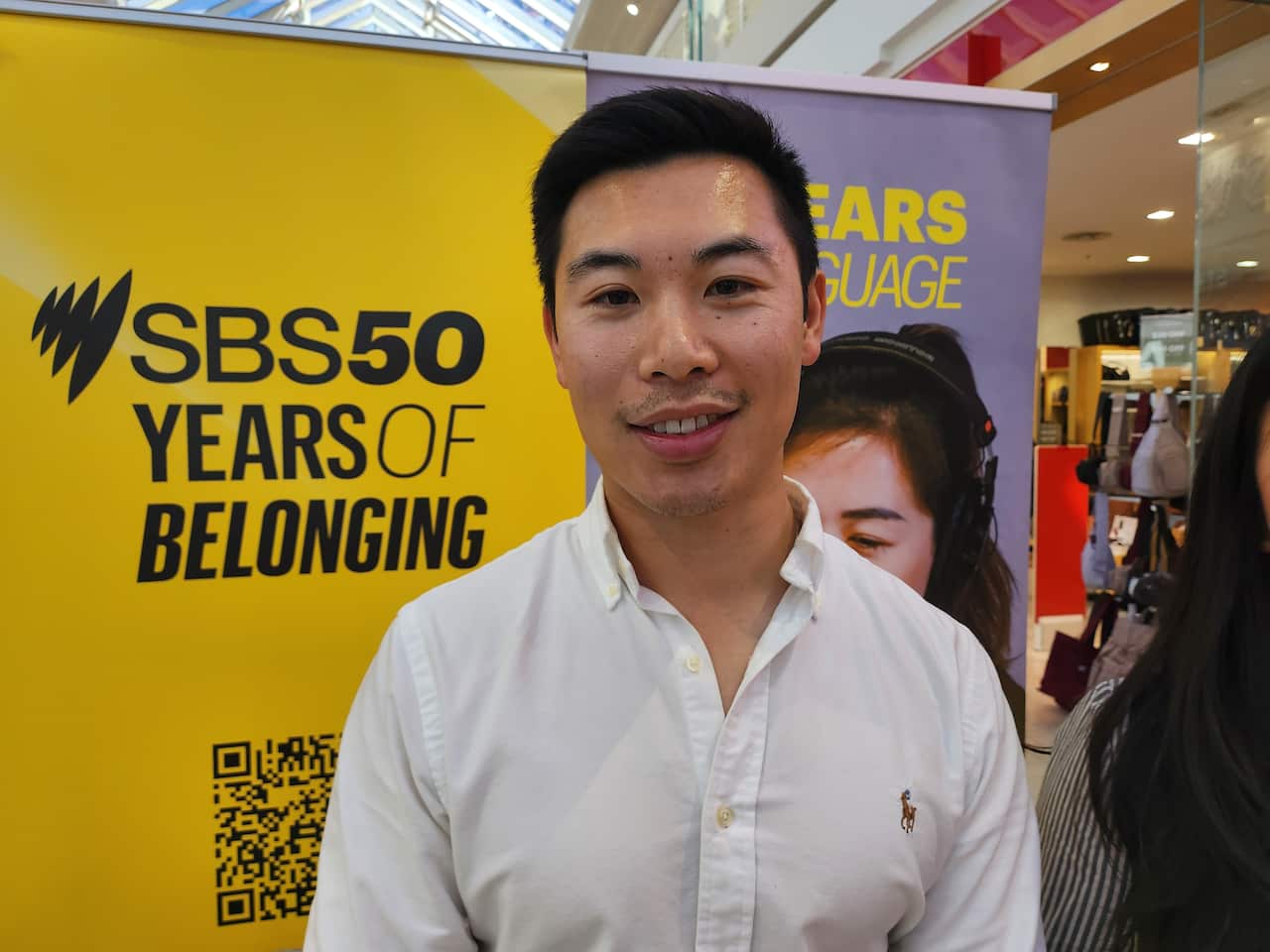 A man in a white shirt smiles in front of a poster that says 'SBS 50 years of belonging'.