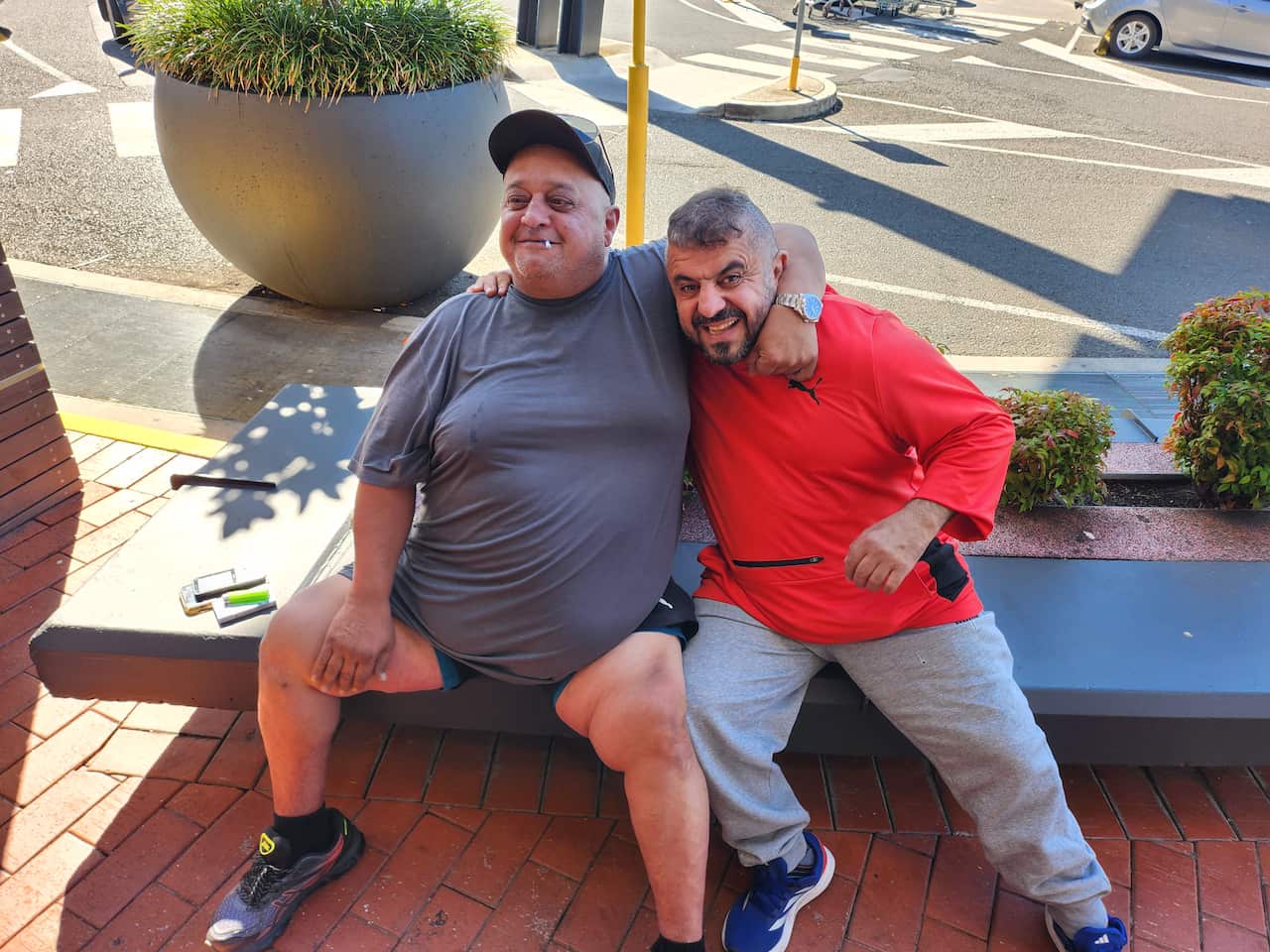 A man in a grey shirt has his arms around a man in a red shirt. They are sitting on a bench.
