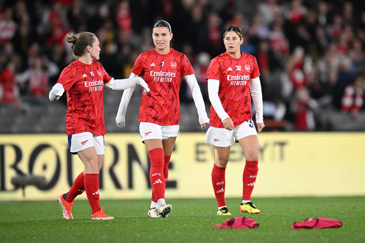 Three players wearing red Arsenal football team jerseys walk across a football field. 