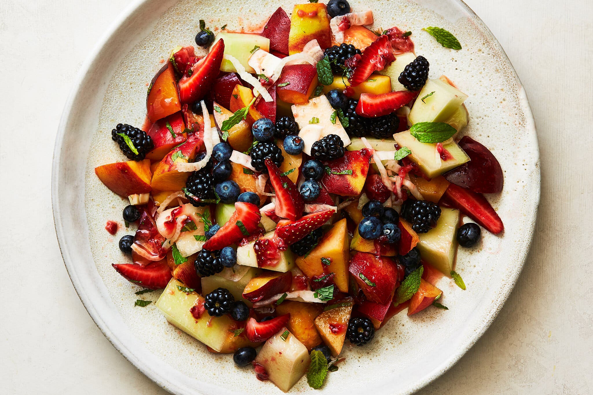 An overhead image of a speckled white plate topped with berries and cubed melon topped in a sauce and sprinkled with herbs.