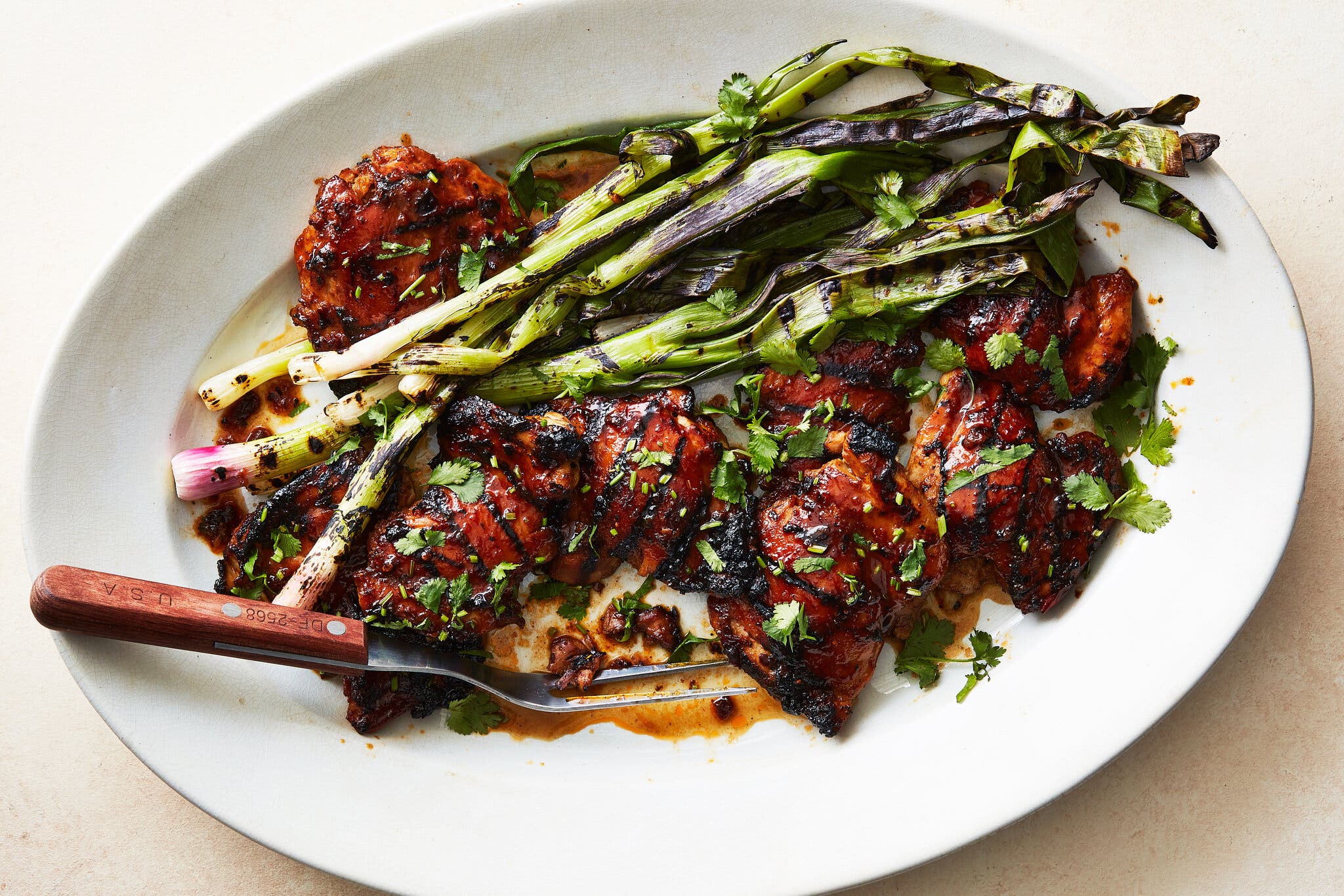 A white platter topped with grill-marked chicken pieces and strips of roasted scallions is photographed from overhead.