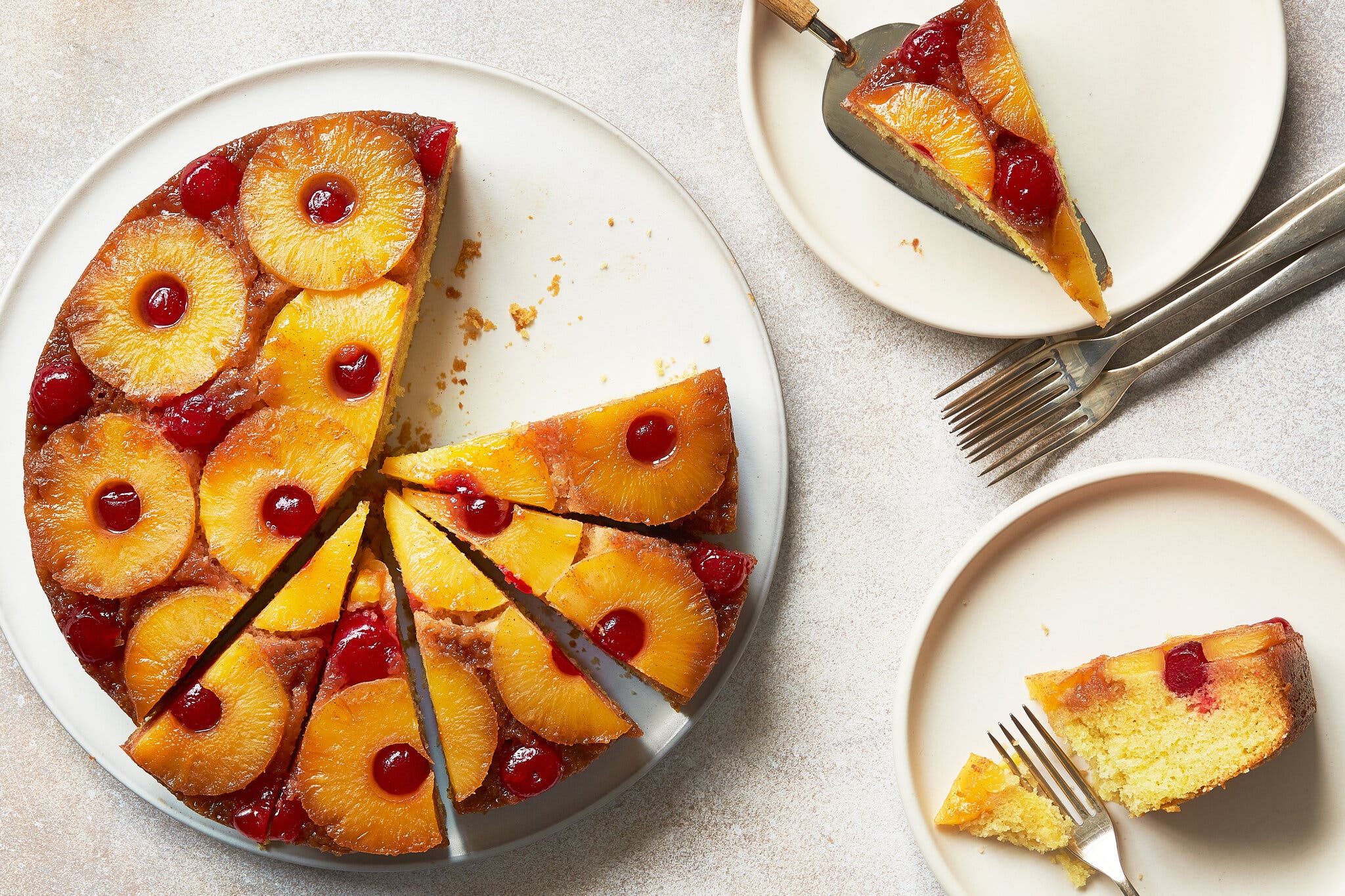 An overhead image of an upside-down cake on a white platter. A good part of it has been sliced, and to the right are two plates with slices and forks.