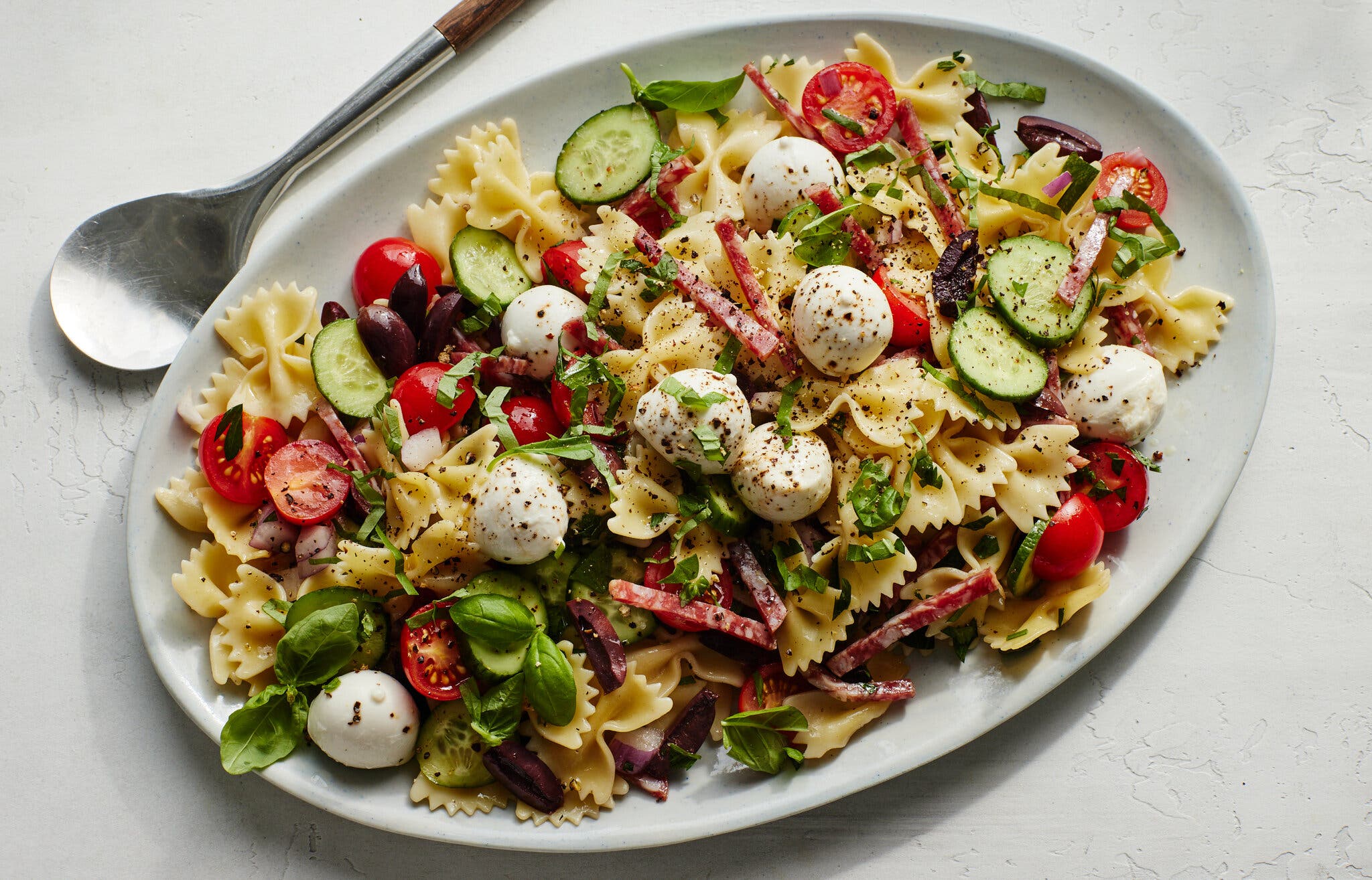 An overhead image of a white platter topped with bowtie pasta, mozzarella balls, sliced olives, tomatoes and cucumber.