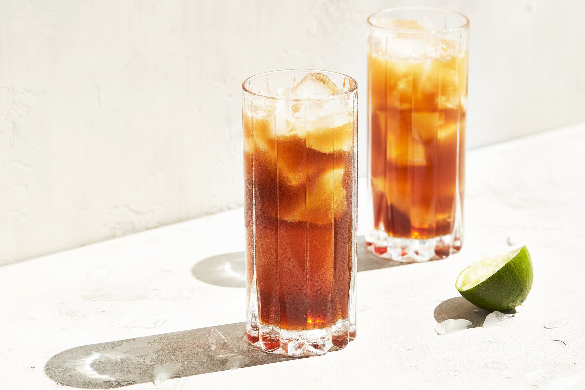 Two clear tall glasses hold ice and an amber liquid. They cast their shadows over a white backdrop. A spent lime half sits just off to the side.