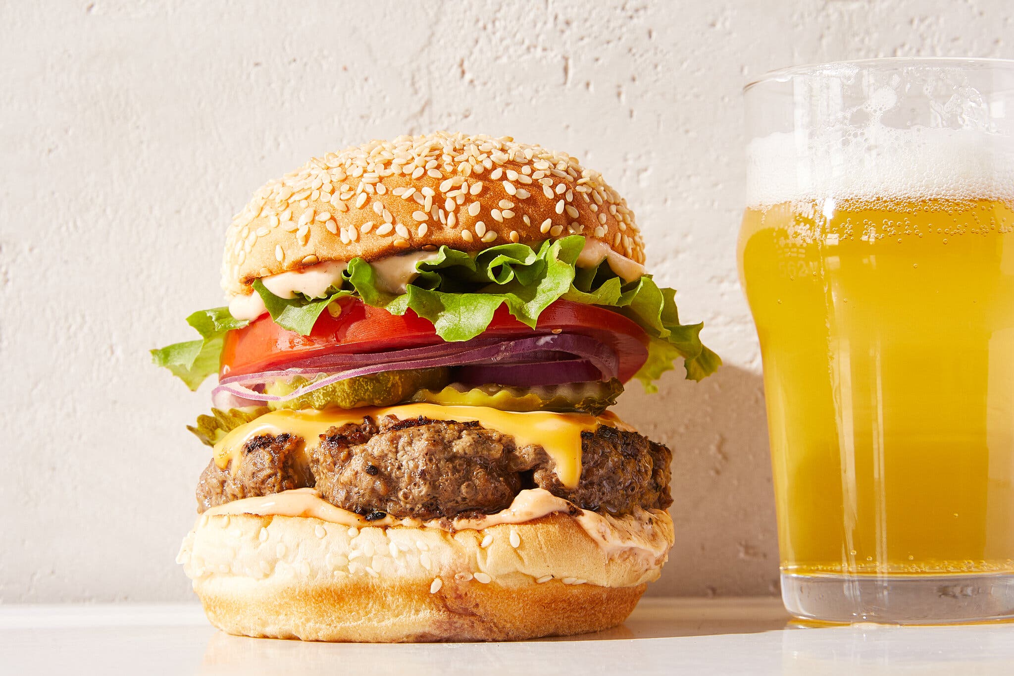A side image of a cheeseburger layered with lettuce, tomato, onion and pickles, and topped with a sesame seed bun. To the right is a pint of beer.