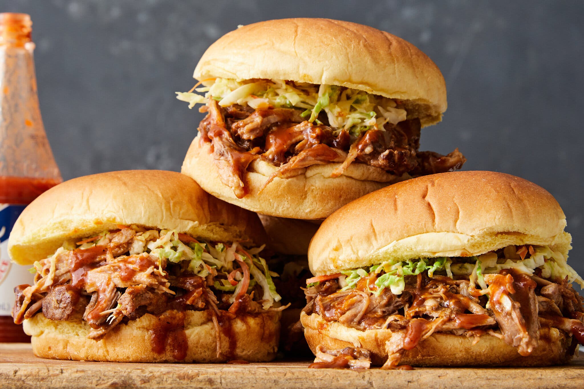 A side image of three pulled pork sandwiches on a cutting board. An open bottle of hot sauce sits in the background.