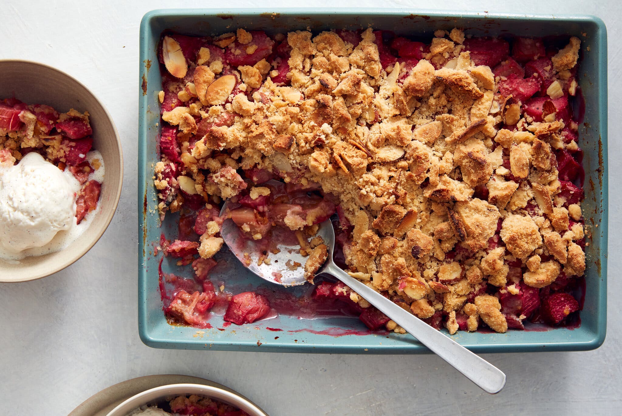 An overhead image of a baking dish filled with chunks of rhubarb and topped with a crisp, browned sweet topping. To the left is a bowl with a serving, topped with ice cream.