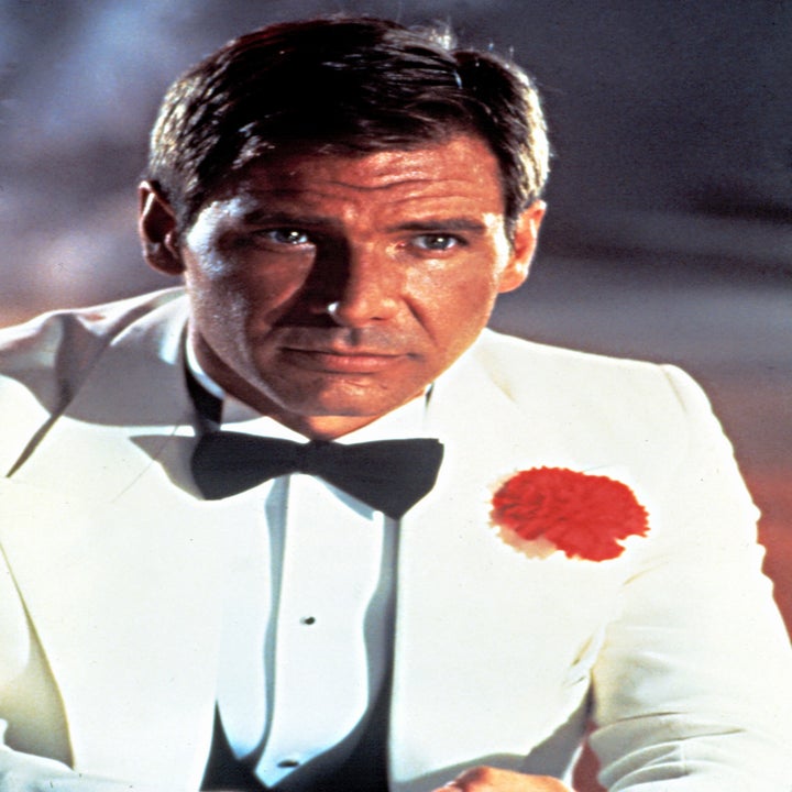 Person in a white tuxedo with a red flower on the lapel, wearing a black bow tie, looks seriously ahead