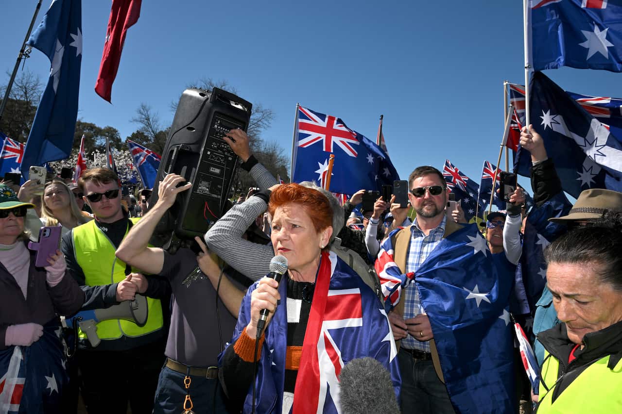 Pauline Hanson wearing an Australian flag, holding a microphone and speaking into it. She's surrounded by peolpe, many of whom are also carrying an Australian flag.