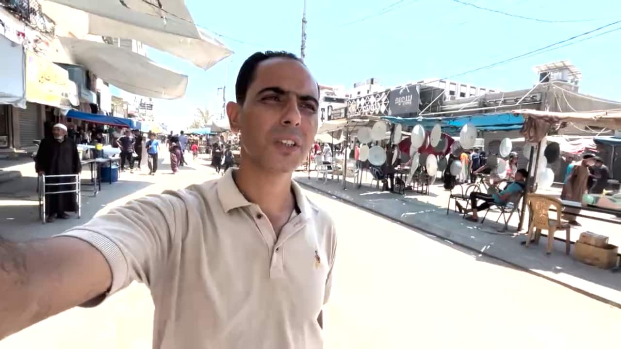 A man in a tan polo shirt stands on a street with market stalls and people in the background.
