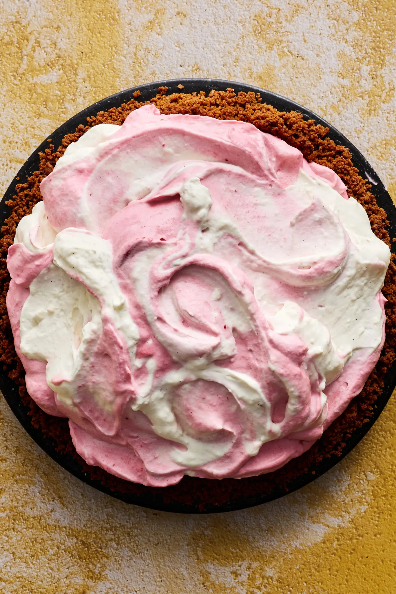 An overhead shot of a pie with a crumbly brown crust and dolloped-in swirled pink and white mouse sitting on a yellow and white background.