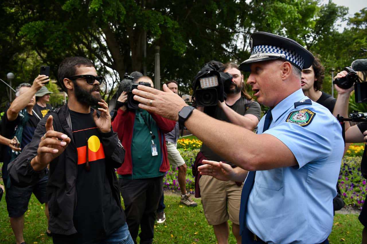 An Australian police officer gestures toward a man wearing an Aboriginal flag shirt in a park, while several members of the media record the interaction.