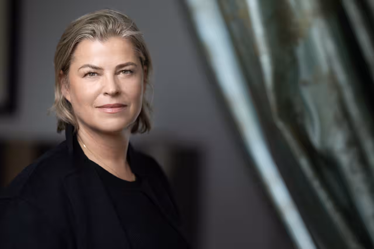 A woman with blonde hair and wearing black clothing stares into the camera 
