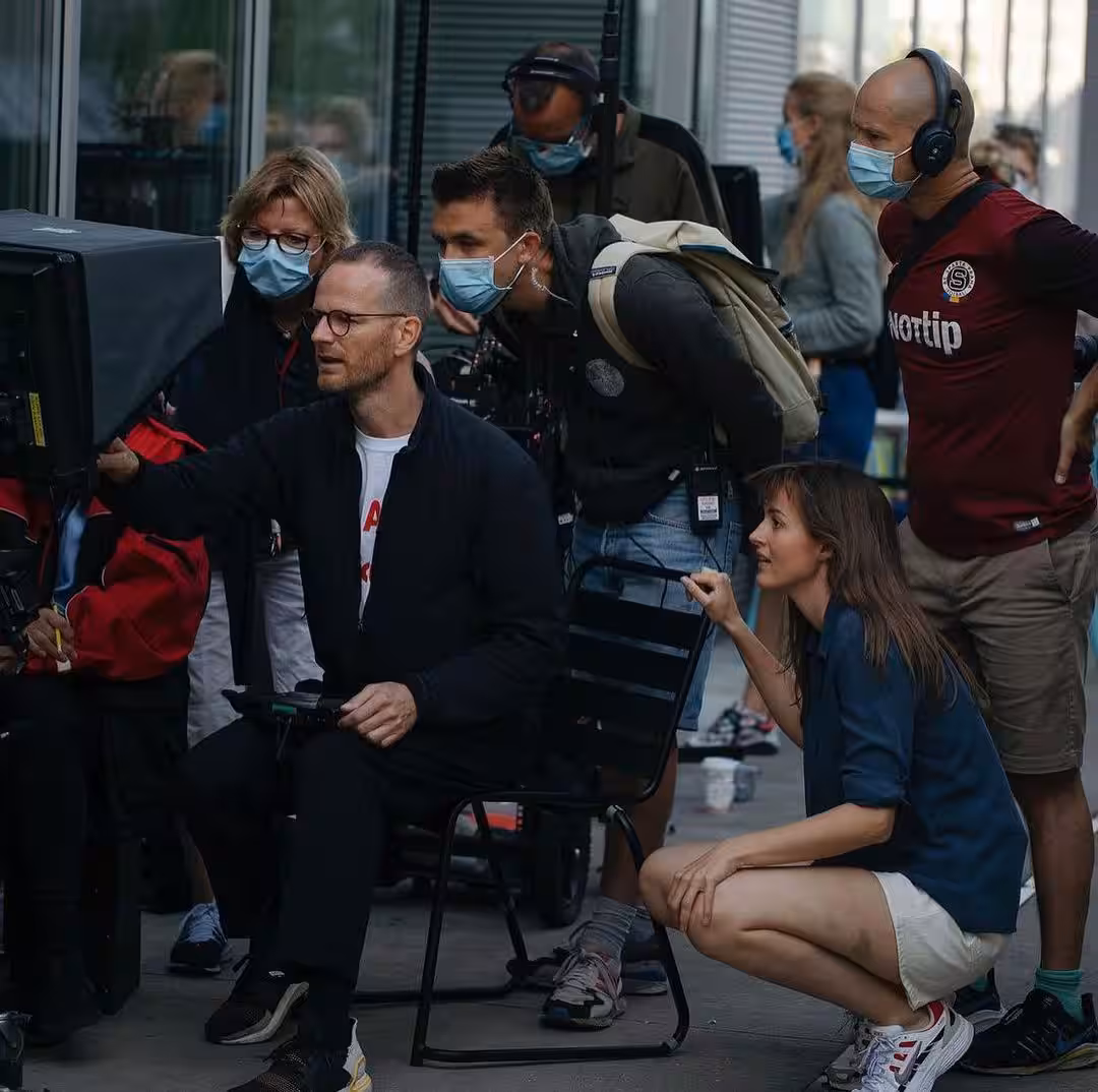 A group of crew members and actors, several wearing face masks, gather around a camera monitor on a film set to review a shot.