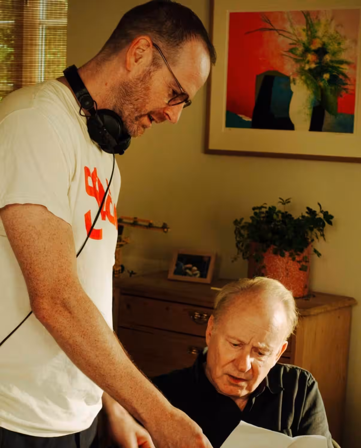 A man wearing headphones leans over an older man to review a document together in a room with warm lighting and a wooden dresser.