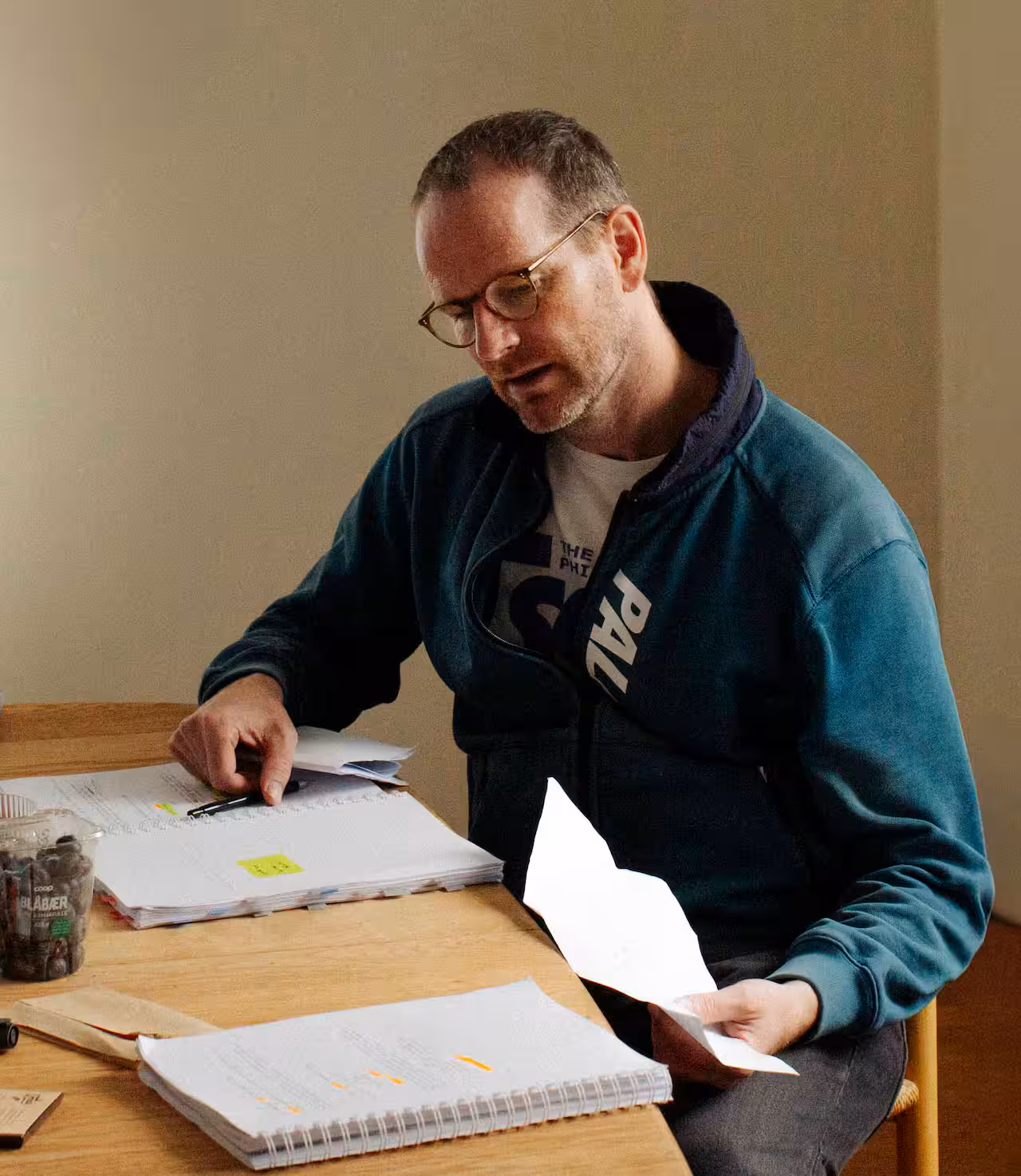 A man wearing glasses and a blue zip-up jacket sits at a table, focused on reviewing a notebook with sticky notes and holding a pen.