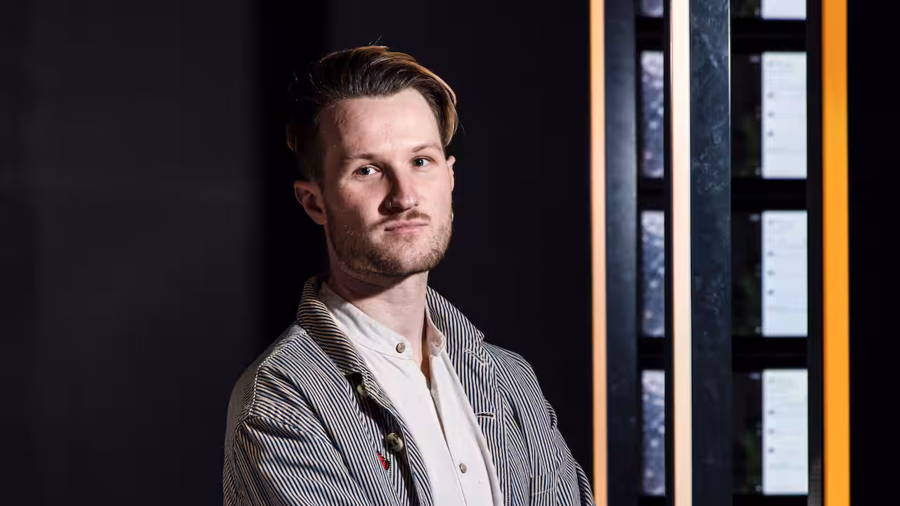 A man with short brown hair, wearing a white shirt and pinstripe jacket. He's staring at the camera.