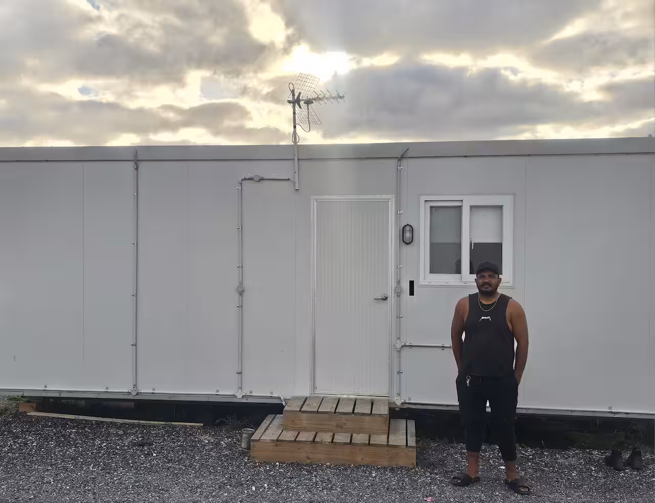 A man wearing a black singlet and trackpants stands outside a white caravan.