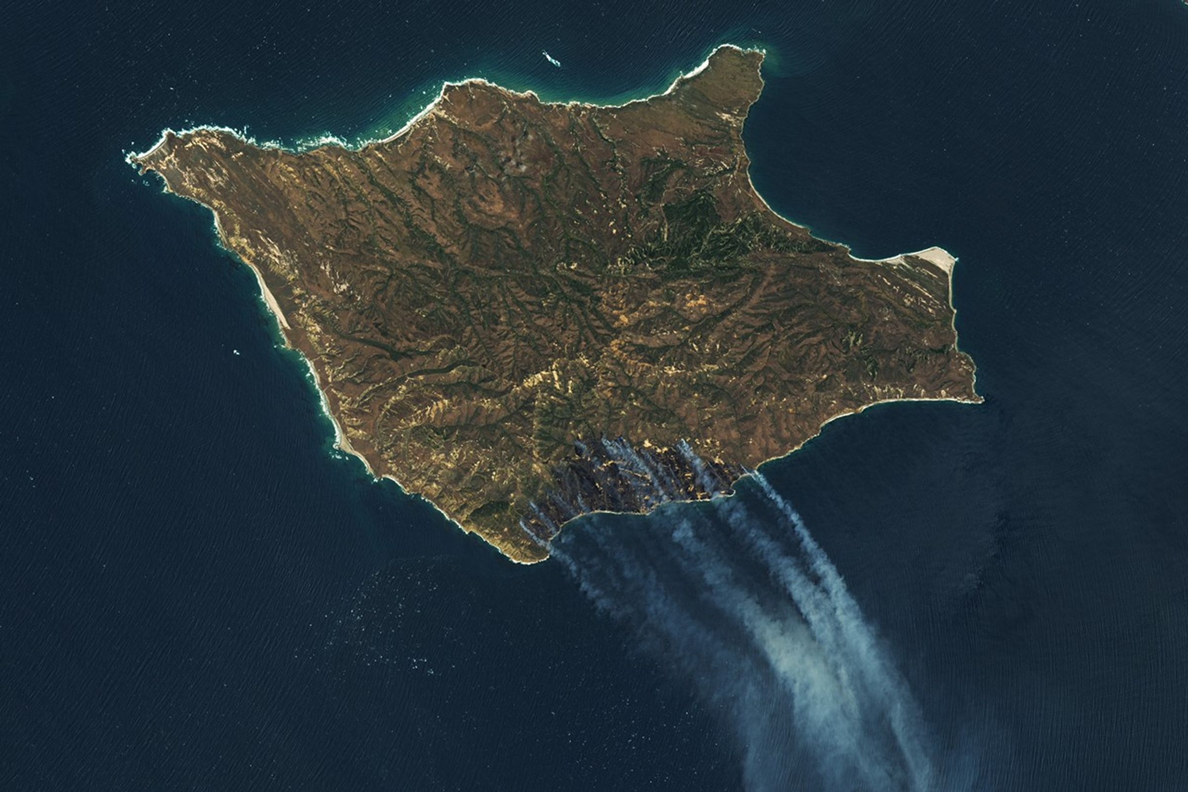 A downward-looking image of Santa Rosa Island is mostly brown, with a darker brown area on the bottom-right side. Gray-white smoke drifts toward the bottom-right over dark blue ocean water.