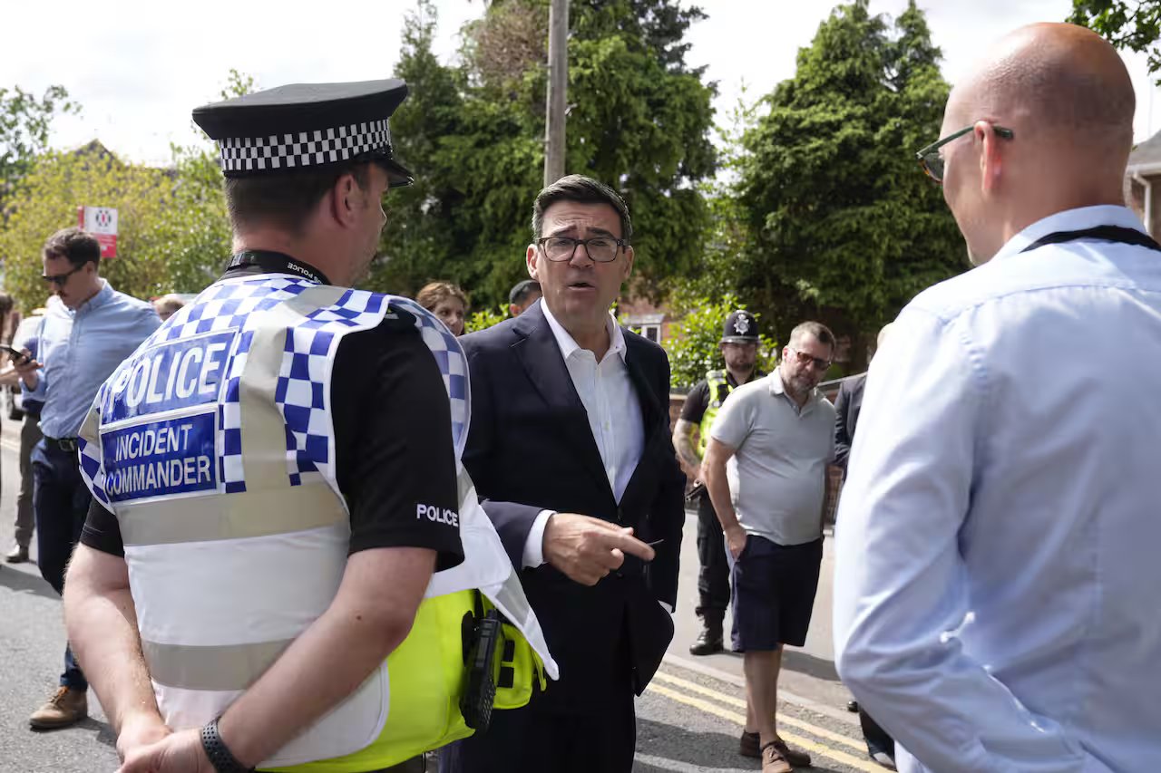 UK PM Andy Burnham in a suit talks to two men in uniform with their backs to the camera.