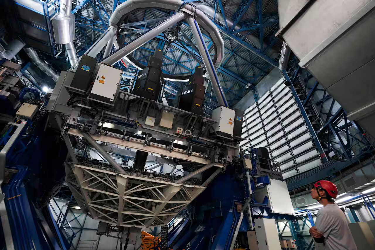 A man wearing a red helmet and grey shirt looks up at a large telescope inside an industrial centre.