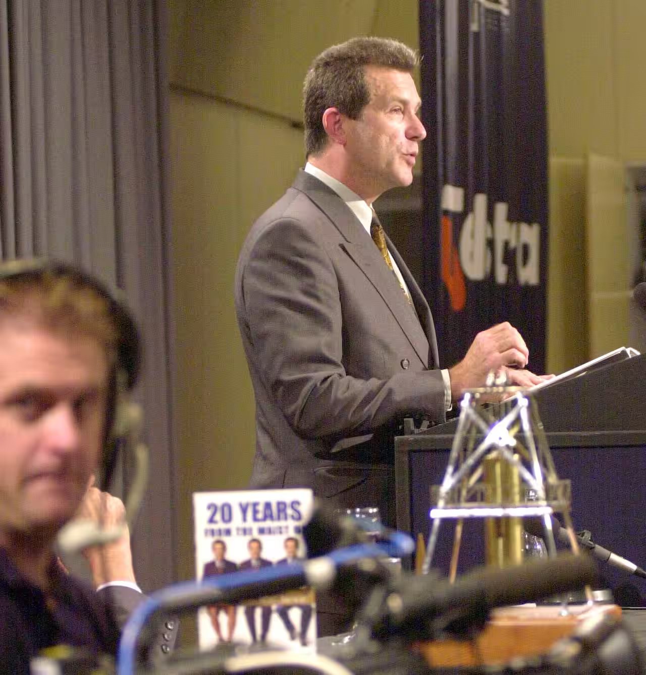 A man in a suit speaks into a microphone while standing at a lectern during a presentation.